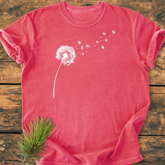 Dandelion Vintage Wash Tee featuring a white dandelion graphic on a red crew-neck, laid flat on wood. Styled with a pine sprig, highlighting its cozy, garment-dyed, relaxed fit.