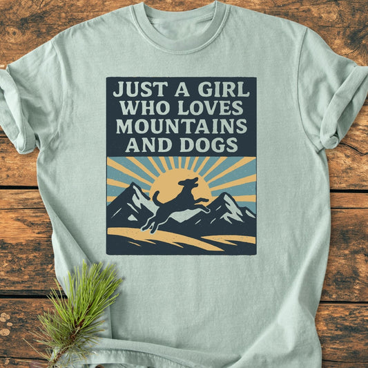 Just A Girl Who Loves Dogs - Vintage Wash Tee; light-teal T-shirt featuring a retro dog and mountain print, laid on a wooden surface with a pine sprig accent.