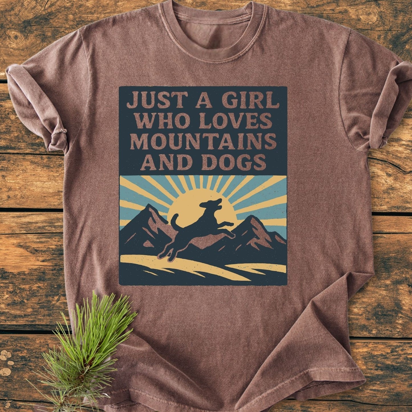 Just A Girl Who Loves Dogs - Vintage Wash Tee features a leaping dog silhouette with mountains and a sun graphic, on a soft, brown T-shirt laid flat on a wooden surface.