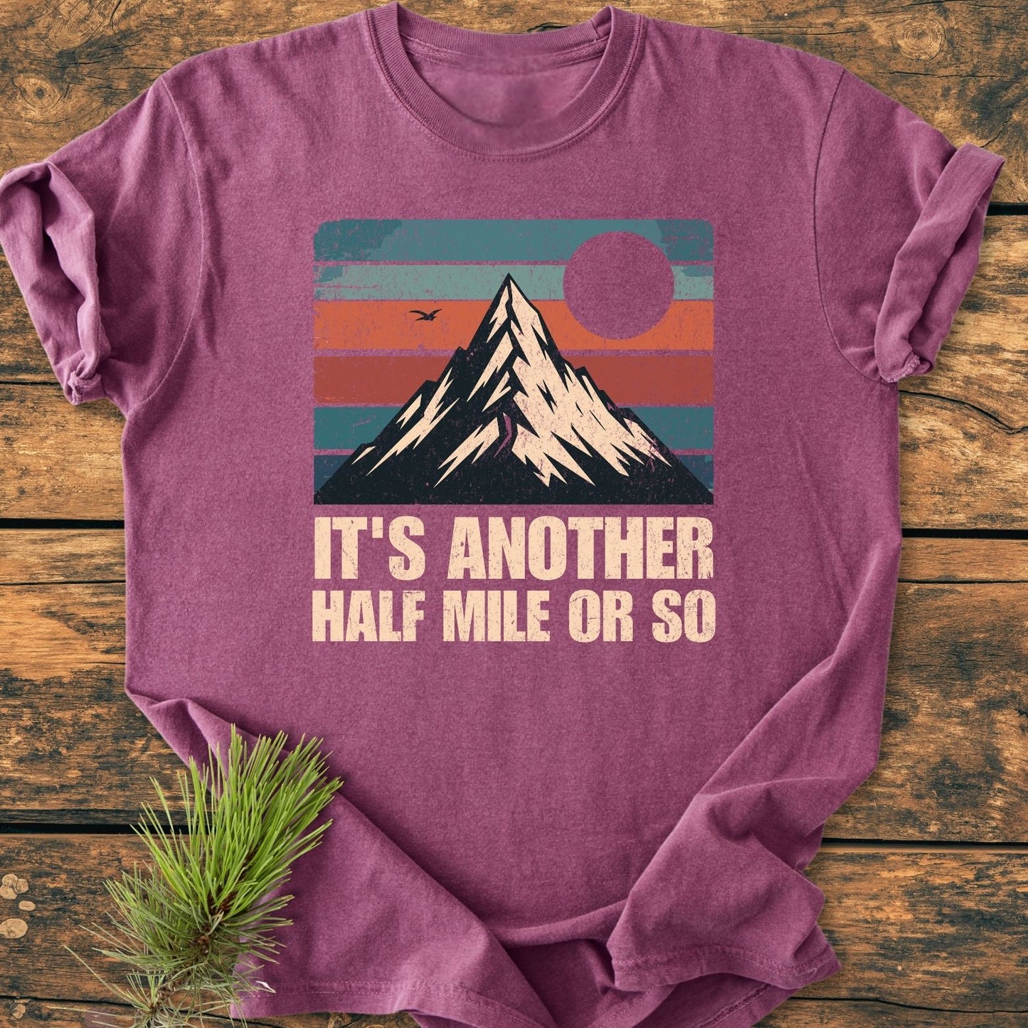 It's Another Half Mile - Vintage Wash Tee features a retro mountain graphic with sunset stripes and slogan. Laid on wood, styled with pine branch. Relaxed fit, soft-washed, durable cotton.