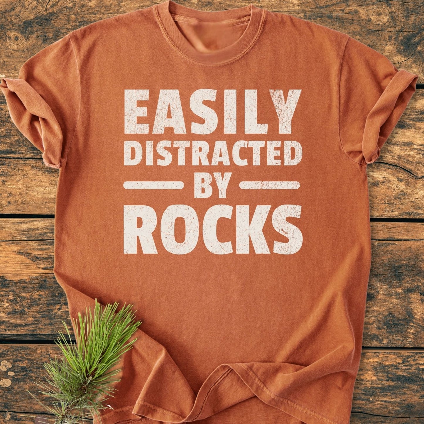 Easily Distracted By Rocks - Vintage Wash Tee, featuring distressed white lettering on a rust-orange cotton shirt. Laid flat on wood with a pine sprig nearby, showcasing its relaxed, durable design.