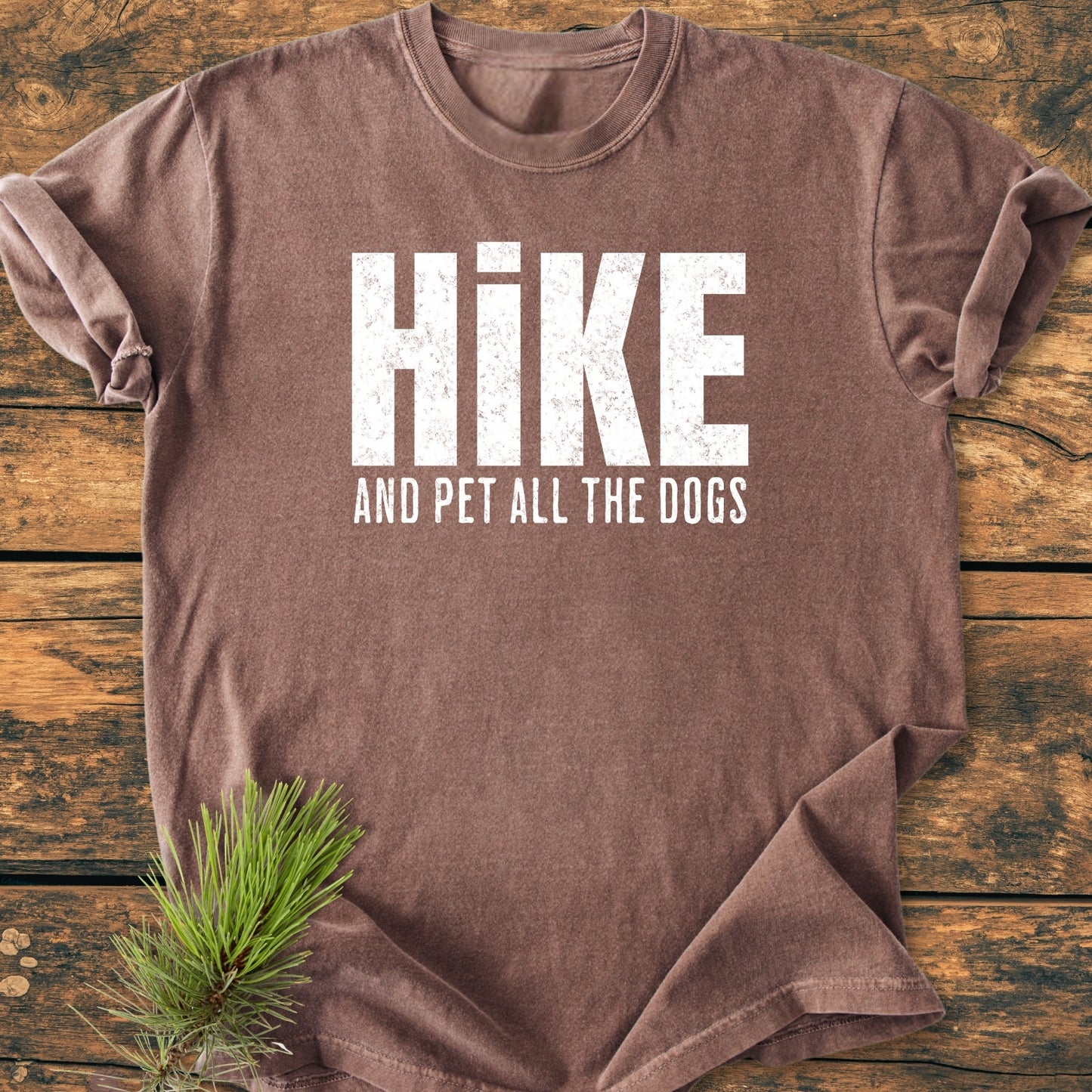 Hike And Pet All The Dogs - Vintage Wash Tee: Brown short-sleeved T-shirt with HIKE and AND PET ALL THE DOGS in white distressed text, laid on a wooden surface with pine sprig.