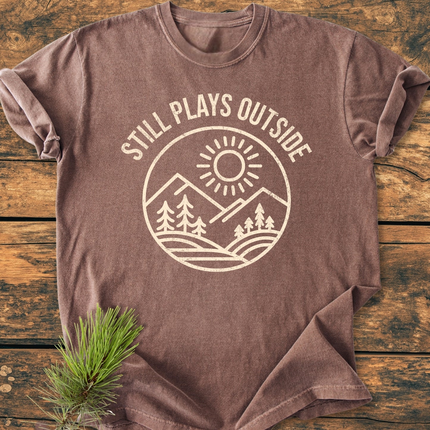 Still Plays Outside - Vintage Wash Tee featuring a cream mountain and sun design on brown, laid on barn-wood surface. Includes pine sprig styling, highlighting the relaxed, durable, garment-dyed cotton fabric.
