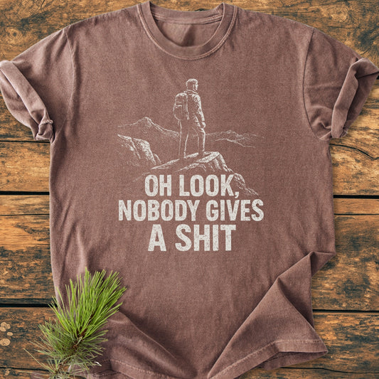 Oh Look - Vintage Wash Tee: Brown short-sleeved T-shirt with white line-art of a backpacker on rocks, bold distressed text, styled with pine sprig on wooden surface.