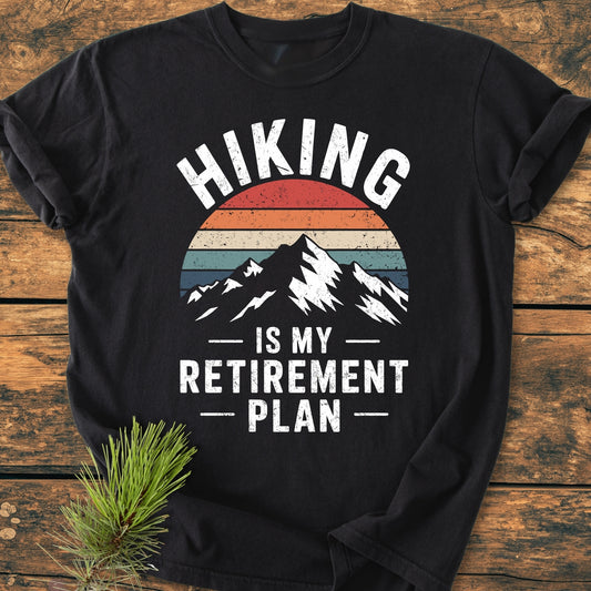 Hiking Is My Retirement Plan - Vintage Wash Tee features a distressed mountain graphic and sunset stripes. Laid flat on wood, this black T-shirt is ideal for outdoor enthusiasts seeking comfort.