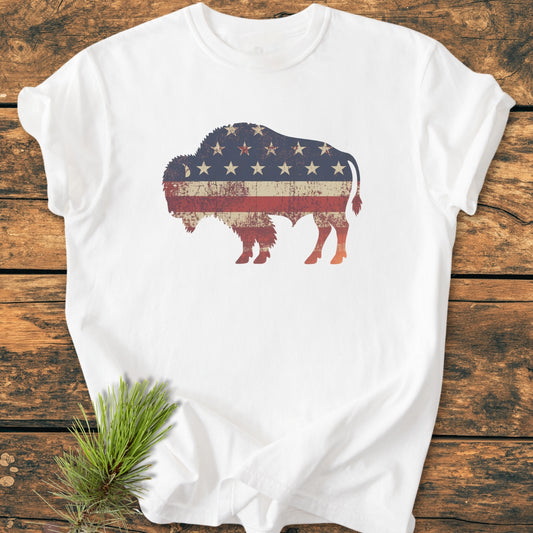 Land Of The Free Bison - Vintage Wash Tee: A white T-shirt featuring a distressed bison silhouette with a vintage U.S. flag motif, laid on rustic wood, accented by a pine sprig.