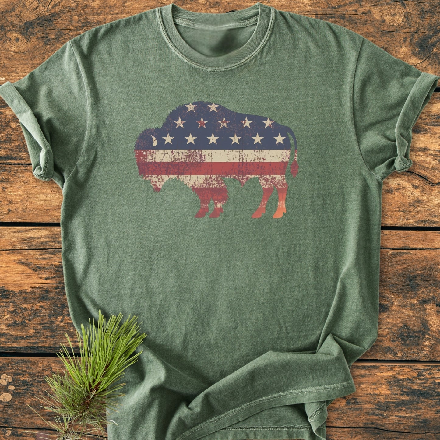 Land Of The Free Bison - Vintage Wash Tee features an olive-green, short-sleeve T-shirt with an American-flag bison silhouette, laid flat on a wooden surface with a pine branch tip visible.