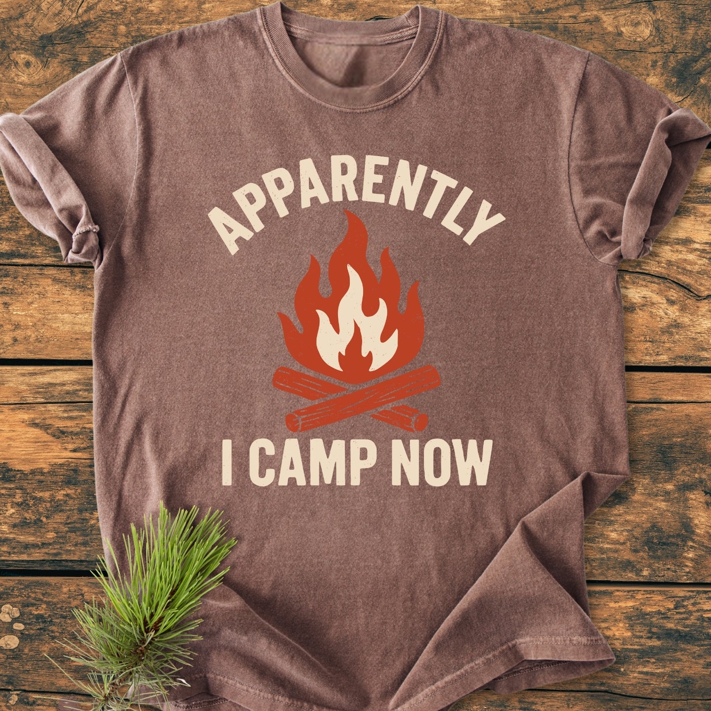 Apparently Camping - Vintage Wash Tee features a stylized campfire graphic and slogan on a brown T-shirt, laid flat with a pine sprig, emphasizing its relaxed, outdoor appeal.