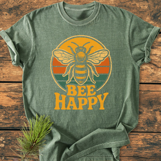 Bee Happy - Vintage Wash Tee in heather-green, featuring a honeybee on a retro striped circle with BEE HAPPY slogan, laid flat on wood with a pine branch accent.