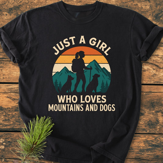 Mountains And Dogs - Vintage Wash Tee with a graphic of mountains, sunset, and a girl hiking with dogs on a black Comfort Colors T-shirt, laid on a wooden surface with pine sprig.