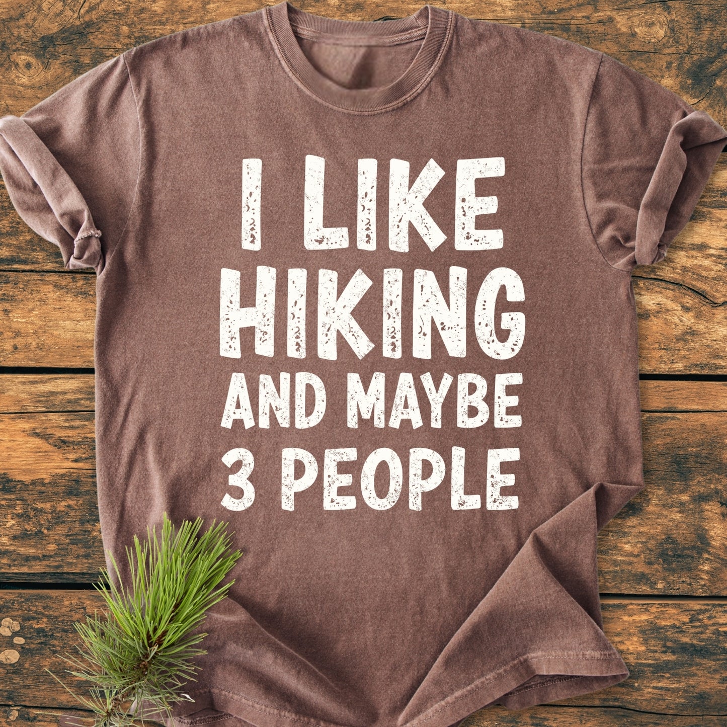 Brown I Like Hiking And Maybe 3 People Vintage Wash Tee with white distressed text, laid flat on wood. Features rolled sleeves and a pine sprig accent, highlighting its relaxed, durable design.