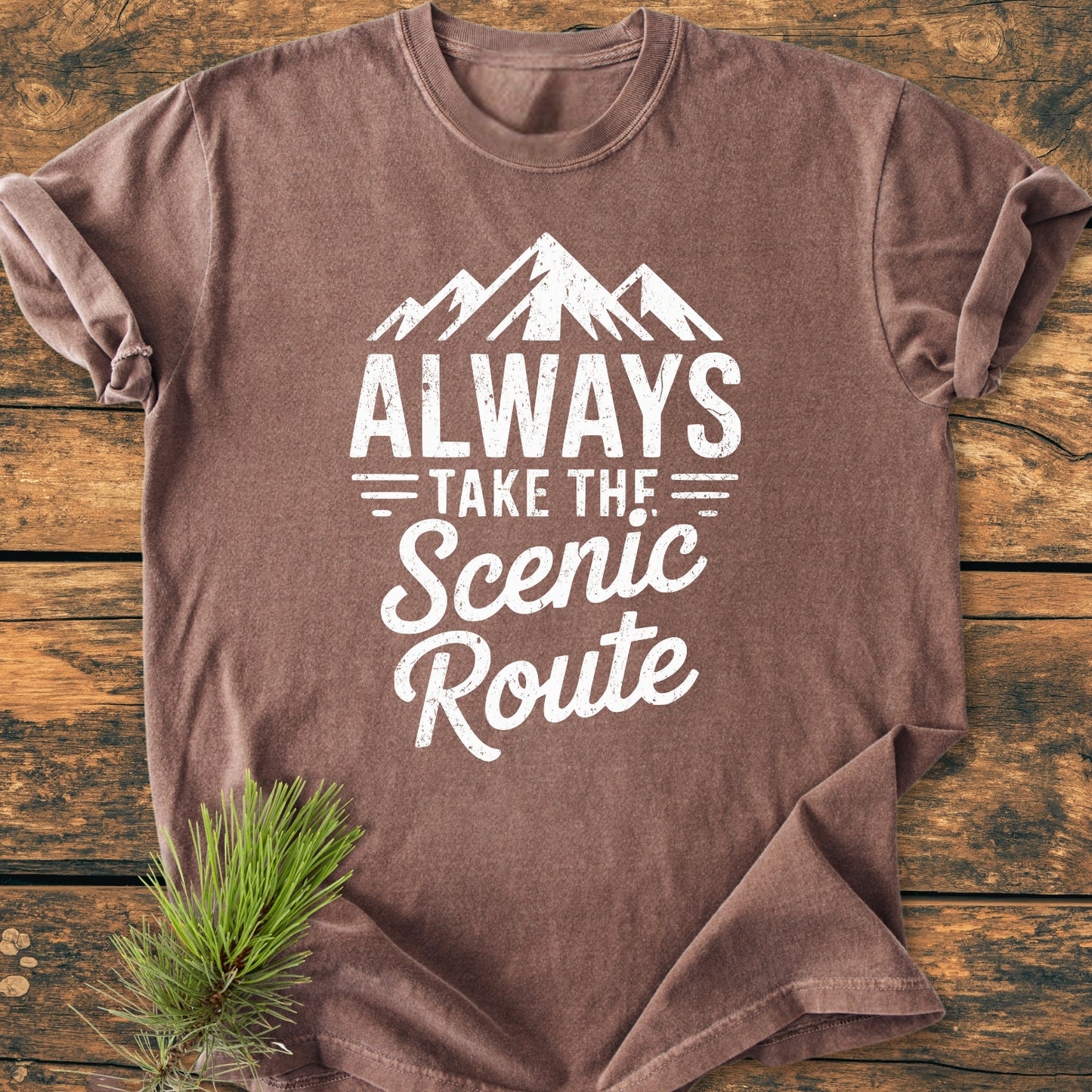 Always Take The Scenic Route - Vintage Wash Tee, featuring a white mountain graphic and slogan on a brown, short-sleeved, soft-washed, garment-dyed fabric, laid on a wooden surface with pine needles nearby.
