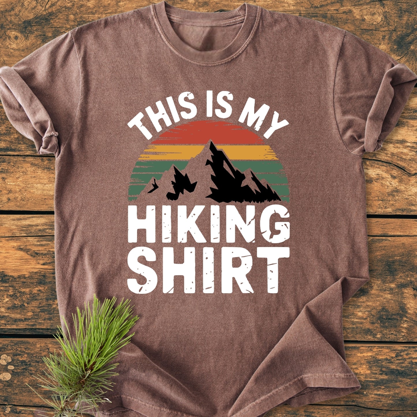 This Is My Hiking Shirt - Vintage Wash Tee features a mountain silhouette against retro sunset stripes with bold text. Laid flat on a wooden surface with a pine sprig beside it.