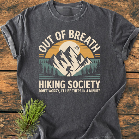 Out Of Breath Hiking Society - Vintage Wash Tee features a mountain range, sun, and hiker print. Displayed on a wooden surface with pine sprig, embodying comfort and durability in a relaxed fit.