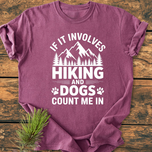 Hiking And Dogs - Vintage Wash Tee featuring a white mountain and paw print graphic on a mauve-purple background, laid flat on wood with a pine needle sprig beside it.