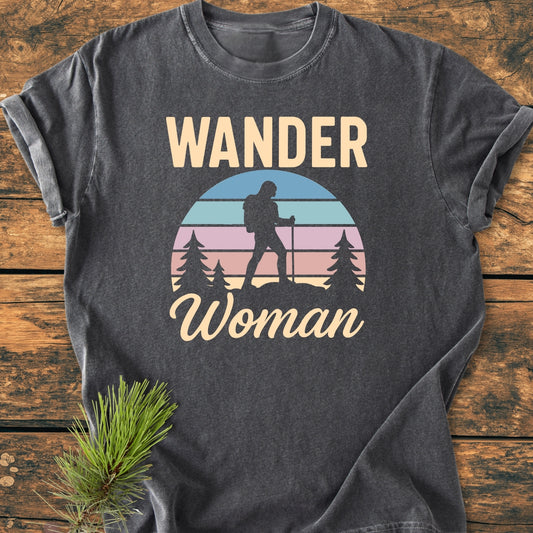 Wander Woman Retro - Vintage Wash Tee featuring WANDER Woman print and pastel sunset graphic, displayed on a wooden surface with a pine sprig. Comfortable, relaxed fit, pre-shrunk 100% ring-spun cotton.