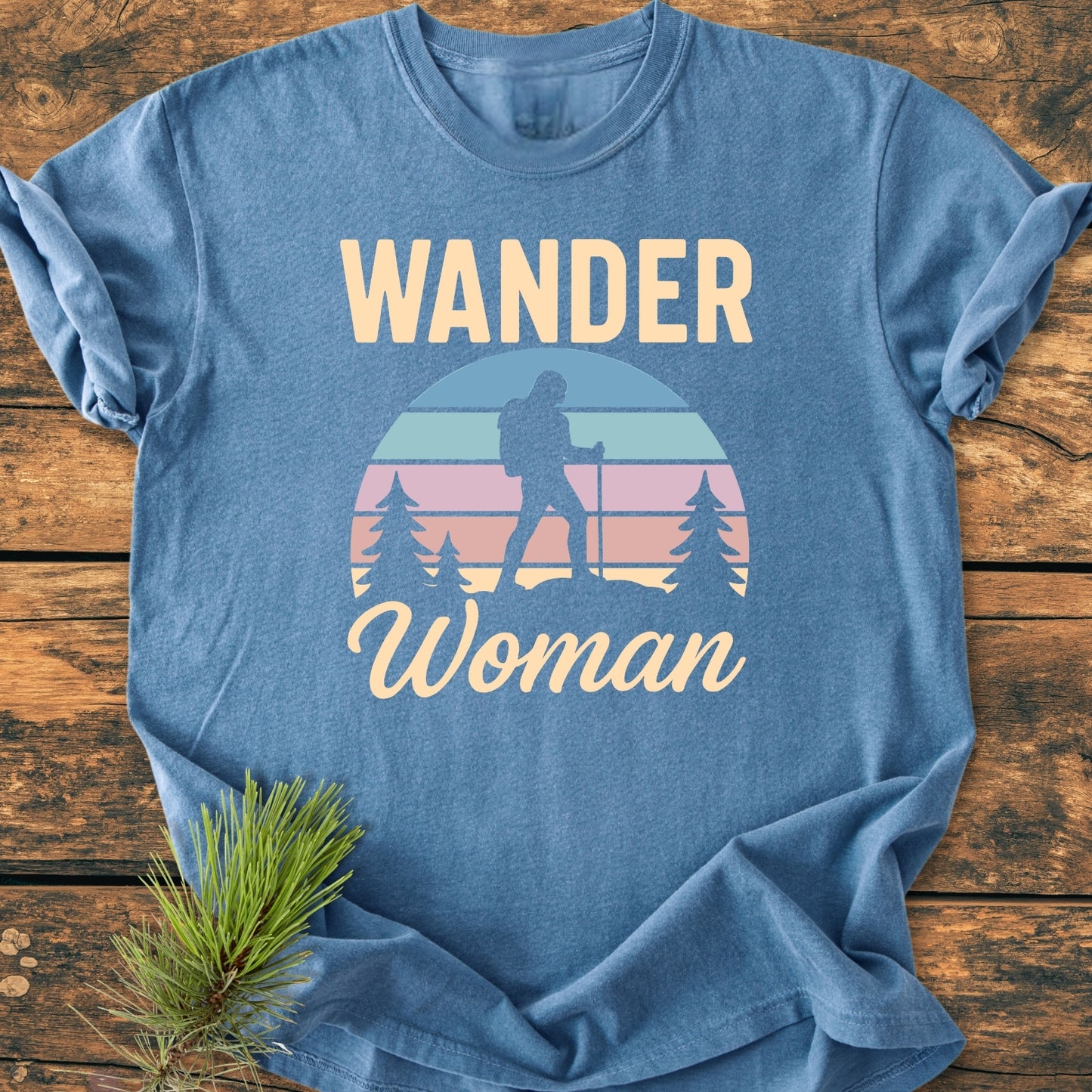 The Wander Woman Retro - Vintage Wash Tee features a heathered-blue design with a female hiker graphic, pine trees, and sunset motif, set on a rustic wooden surface with an evergreen sprig accent.