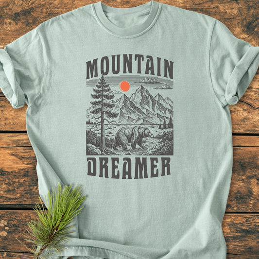 Mountain Dreamer - Vintage Wash Tee featuring a wood-cut outdoor scene with a bear, pine tree, mountains, and red sun. Laid on wooden surface with pine sprig styling.