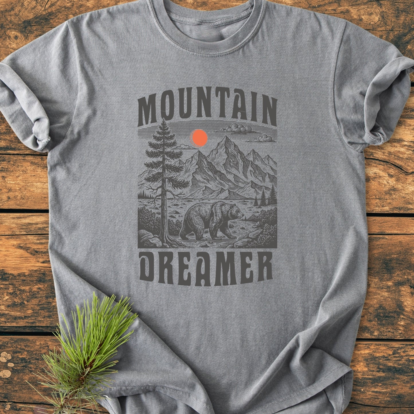 Mountain Dreamer - Vintage Wash Tee features a stylized outdoor scene with pine tree, mountain peaks, sun or moon, and bear, laid flat on rustic wood with pine needles beside it.