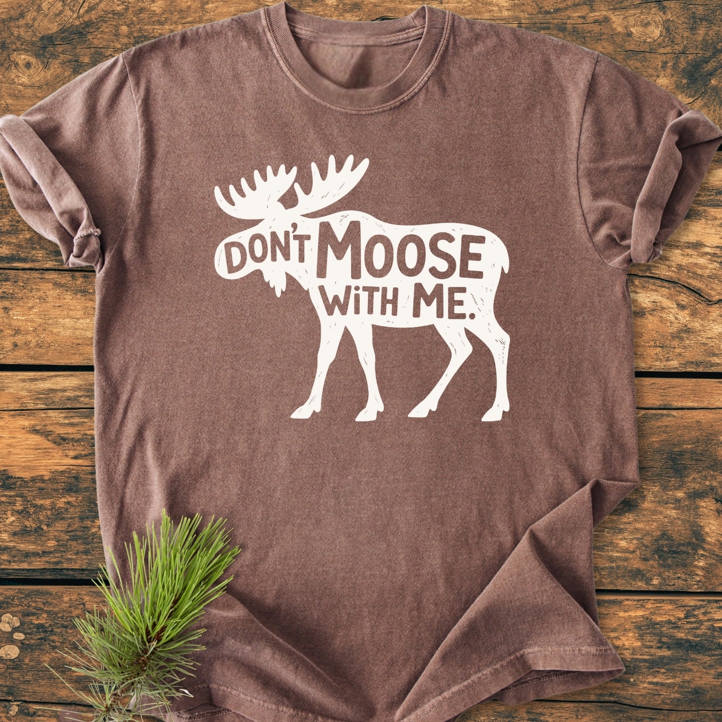 Don't Moose With Me - Vintage Wash Tee featuring a moose silhouette on a heathered brown T-shirt, laid on wood. Accentuated with a pine sprig for a rustic, woodland charm.