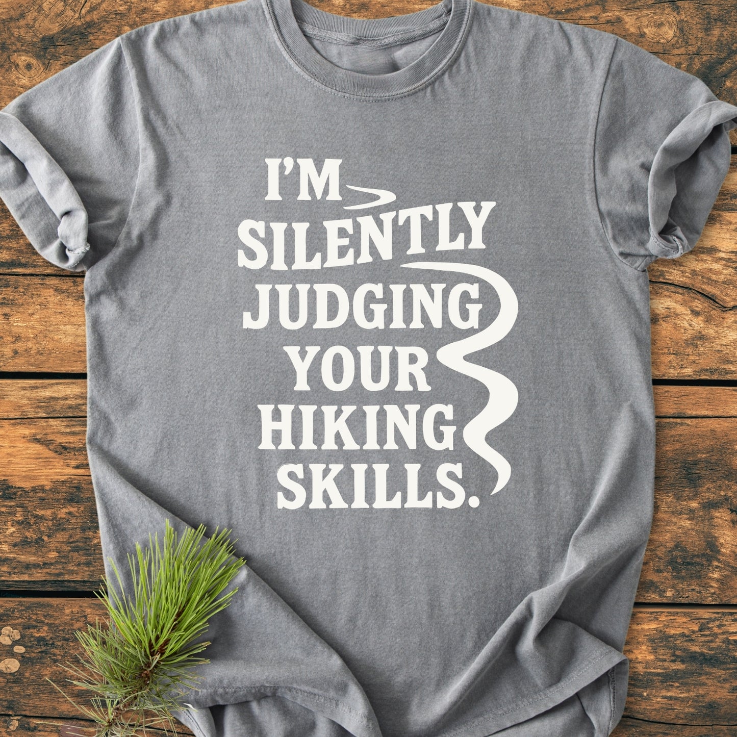 Silently Judging - Vintage Wash Tee in heather-gray, laid flat with slogan in white text and a winding trail graphic, placed on a wooden surface with a pine sprig accent.
