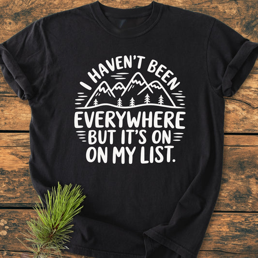 On My List - Vintage Wash Tee featuring stylized mountains and pine trees graphic with travel quote, displayed on a wooden surface, accompanied by a sprig of evergreen for styling.