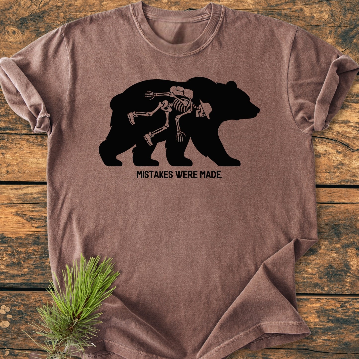 Mistakes Were Made - Vintage Wash Tee featuring a bear silhouette with a hiker skeleton inside, captioned “MISTAKES WERE MADE.” Laid flat on a wooden surface, showcasing its relaxed fit and durable stitching.