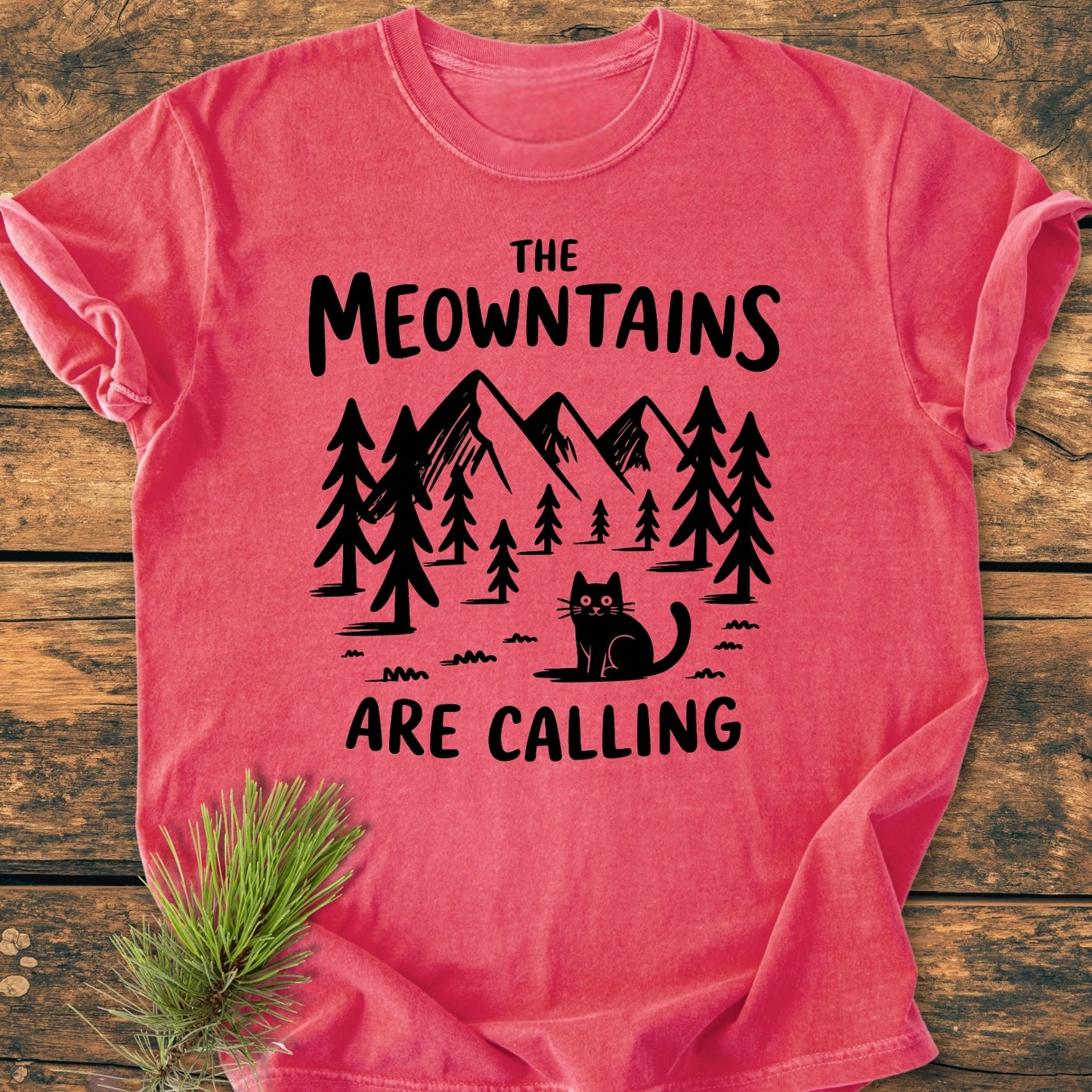 The Meowntains Are Calling - Vintage Wash Tee features a black pine trees and mountains graphic with a cat. Laid on wood, it showcases a punny slogan and includes a nearby pine sprig.