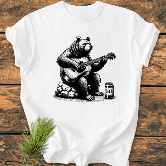 Bear Playing Guitar - Vintage Wash Tee: A white T-shirt featuring a bear playing guitar illustration, with a BEER can nearby, laid on wood with a spruce branch accent.