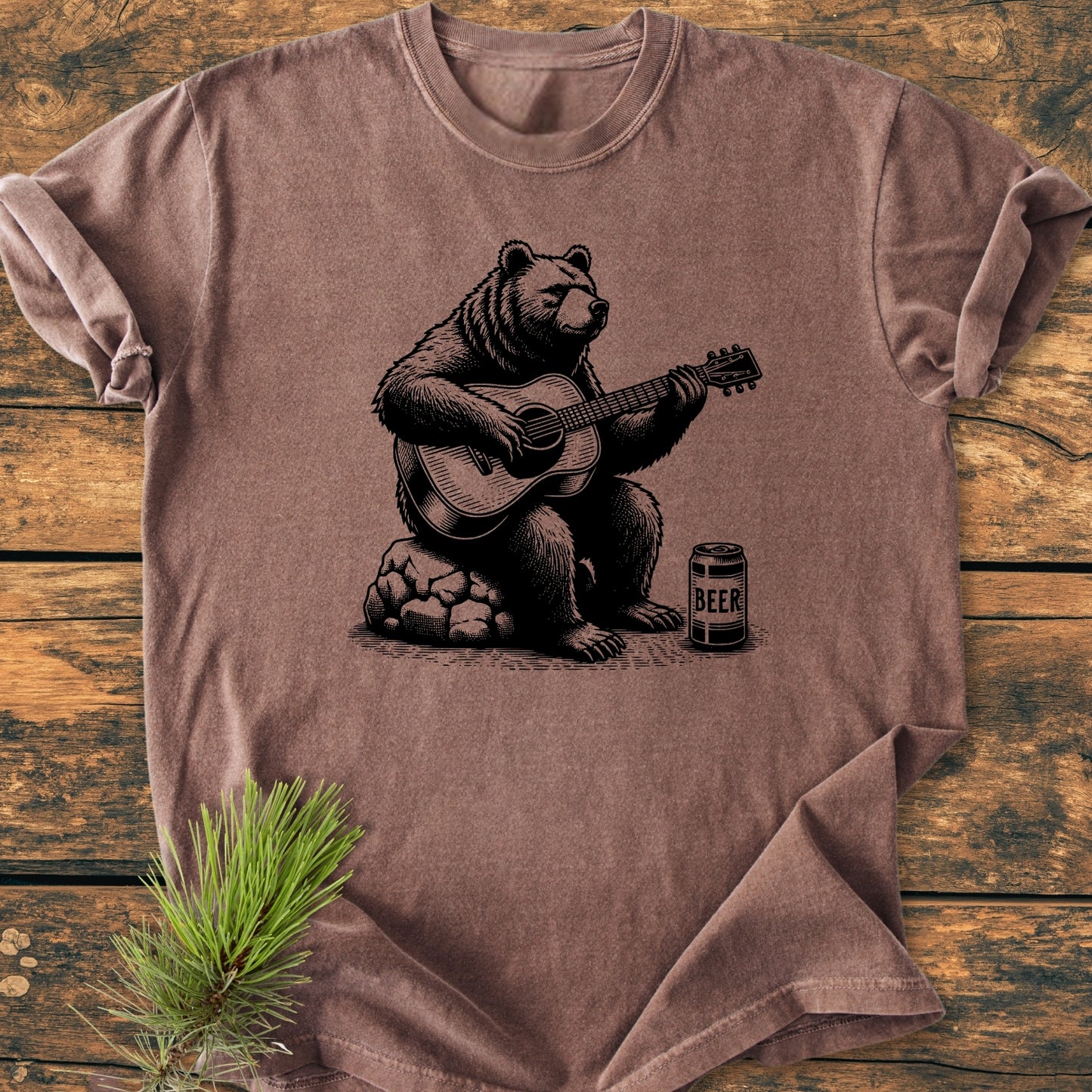 Bear Playing Guitar - Vintage Wash Tee displayed on wooden surface, featuring a bear strumming a guitar beside a BEER can. Includes a pine branch accent, emphasizing rustic charm and relaxed fit.