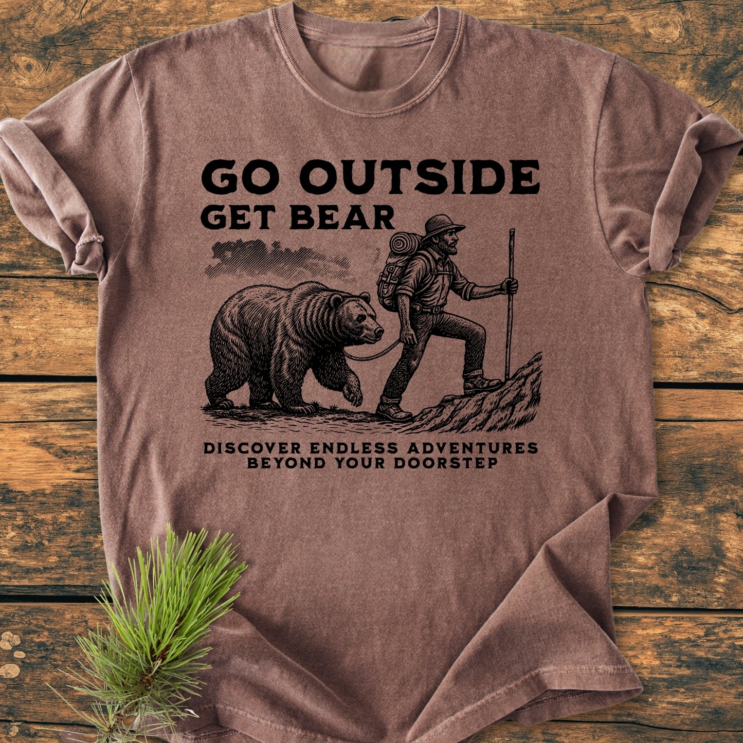 Go Outside Get Bear - Vintage Wash Tee on wooden surface, featuring a hiker with a bear illustration and adventure-themed text, showcasing a comfy, durable design with soft-washed, garment-dyed fabric.