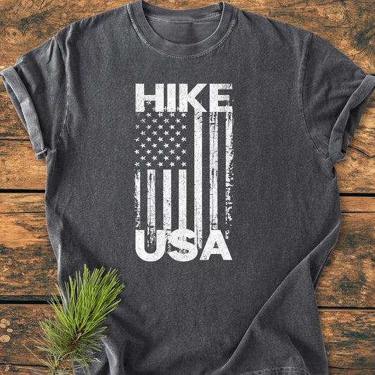 Hike USA - Vintage Wash Tee: A dark gray T-shirt with a worn white vertical American flag graphic, HIKE above, USA in stripes, laid flat on wood, complemented by a small evergreen sprig.