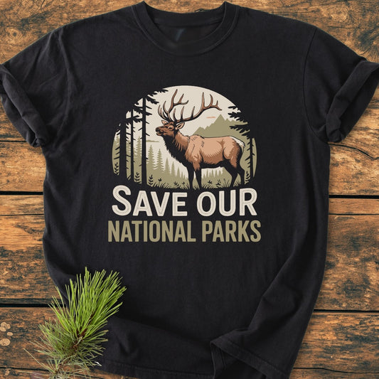 Save Our National Parks - Vintage Wash Tee features a nature-themed elk graphic against trees and mountains, on a black garment-dyed, soft-washed T-shirt, laid on wood with a pine sprig.