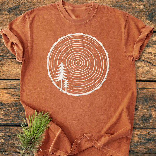 Minimalist Log - Vintage Wash Tee displayed on wood showcases a white tree trunk cross-section and pine tree graphic, emphasizing its rustic appeal and relaxed fit. Pine sprig accent enhances nature-inspired design.