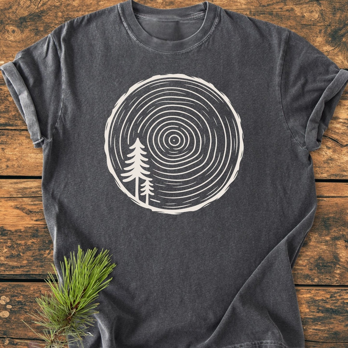 Minimalist Log - Vintage Wash Tee featuring a white tree-ring graphic with pine silhouettes, displayed on a wooden surface, paired with a pine branch. Made from soft, durable, garment-dyed 100% ring-spun cotton.