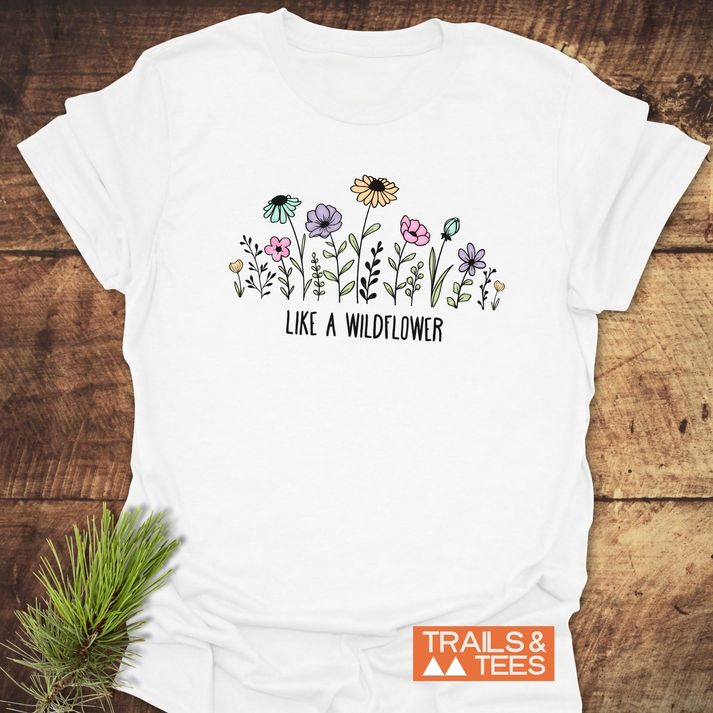 The Like A Wildflower T-Shirt features pastel wildflowers and text on soft cotton, ideal for adventures. Styled with a pine sprig, it's marked by the TRAILS & TEES logo.