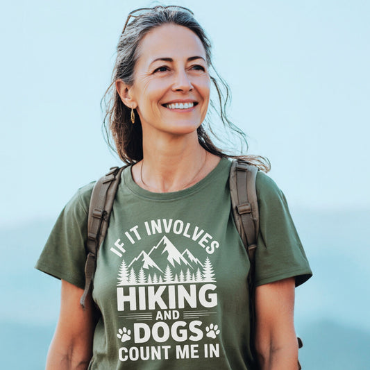 Middle-aged woman wearing the Hiking And Dogs T-Shirt, featuring mountain and pine graphics, with If it involves hiking and dogs count me in text, standing outdoors with a backpack.