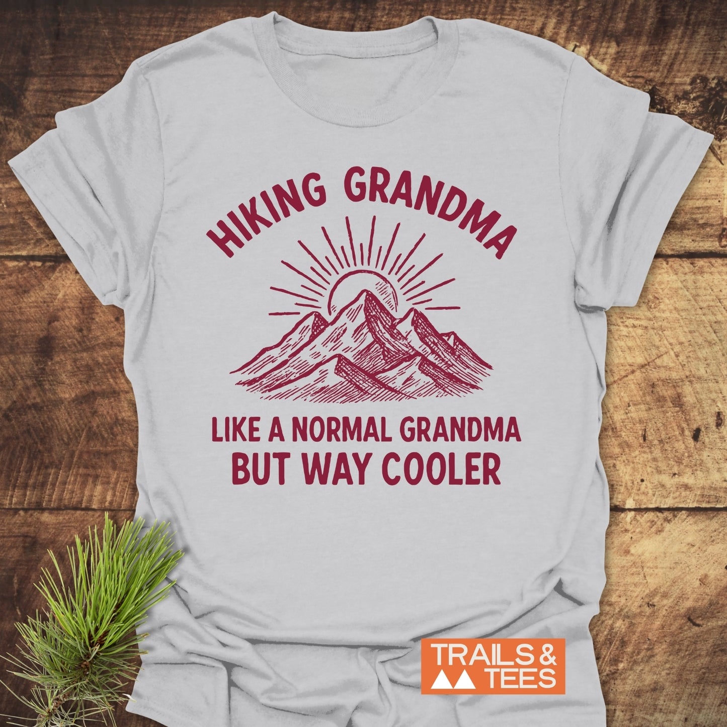 Cool Hiking Grandma Sunrise T-Shirt features mountain peaks with a rising sun graphic, bold red HIKING GRANDMA text, and playful text below. Ideal for outdoor-loving grandmas.