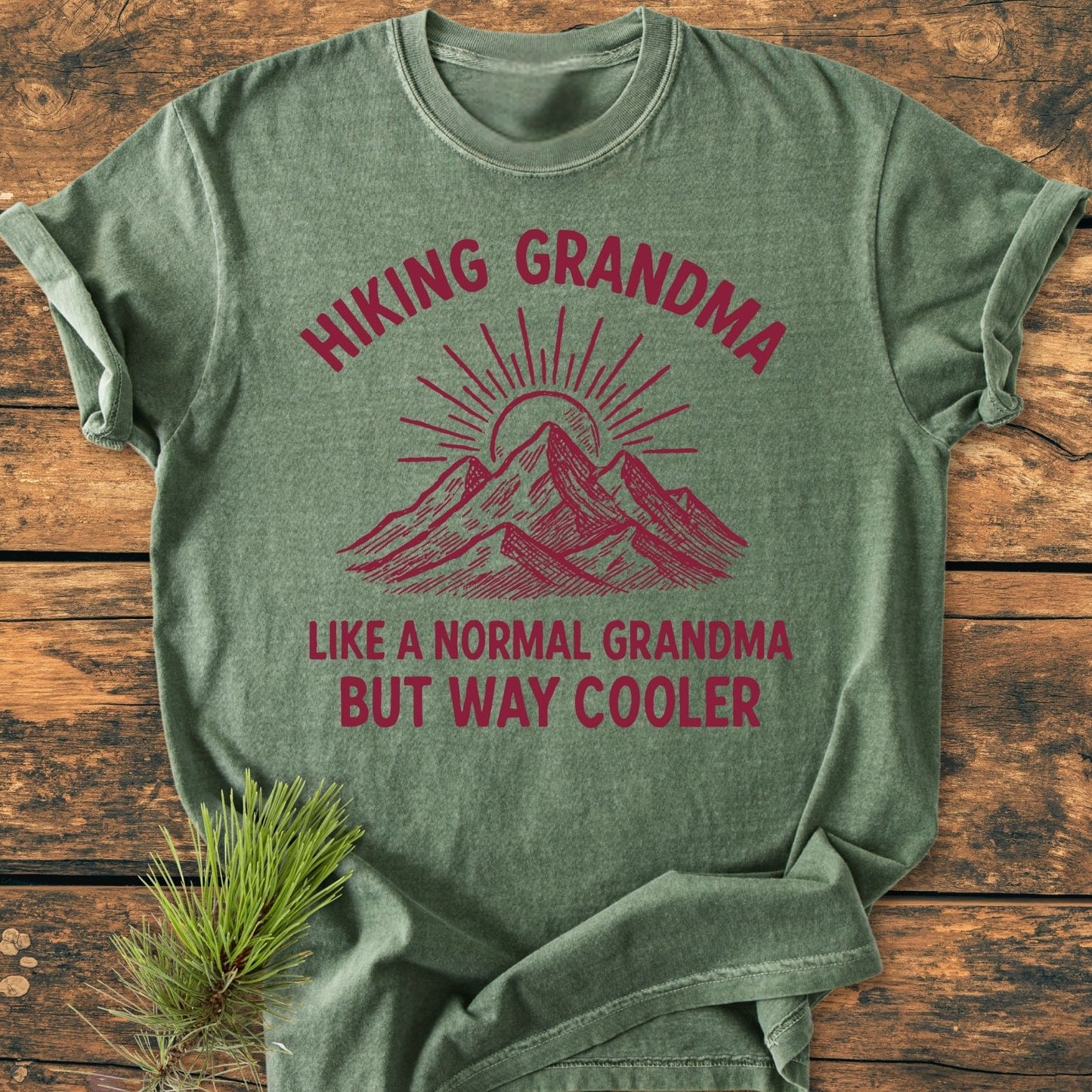 Cool Hiking Grandma Sunrise - Vintage Wash Tee laid flat on rustic wood, features reddish mountain and sun graphic with slogans, and a pine sprig accent. Comfortable and durable, ideal for adventurous grandmas.