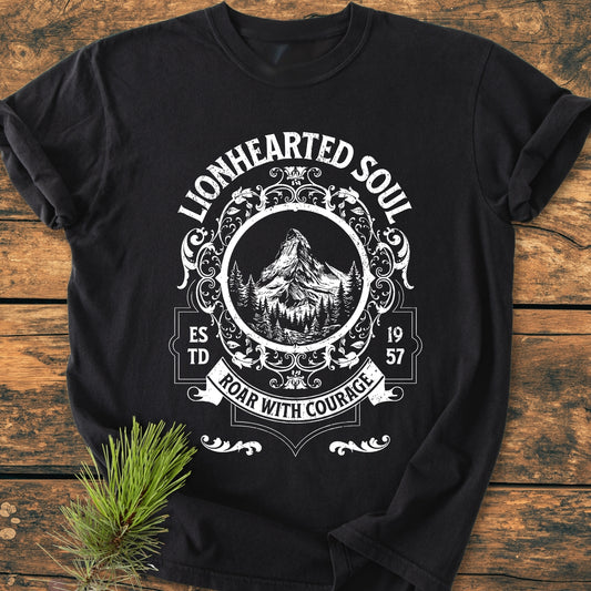 Lionhearted Soul - Vintage Wash Tee features a white, vintage-style emblem with mountains and pine trees on a black T-shirt, laid flat on a rustic wood surface with a nearby pine sprig.