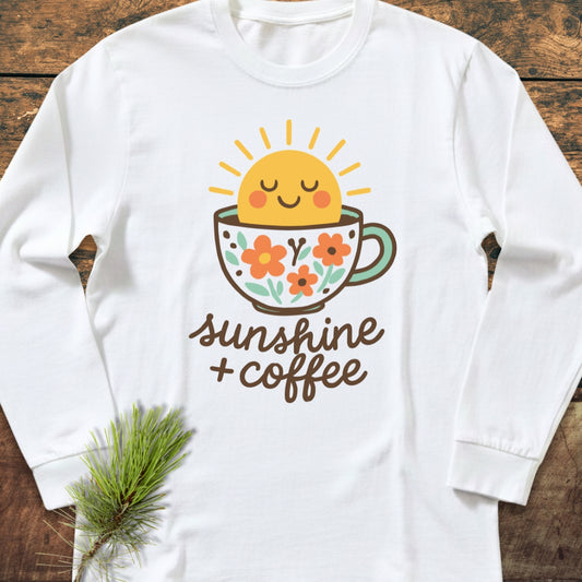 Sunshine And Coffee Long-Sleeve Tee featuring a smiling sun in a flower-patterned cup with sunshine + coffee text, laid on wood with a pine sprig, made of soft ring-spun cotton.
