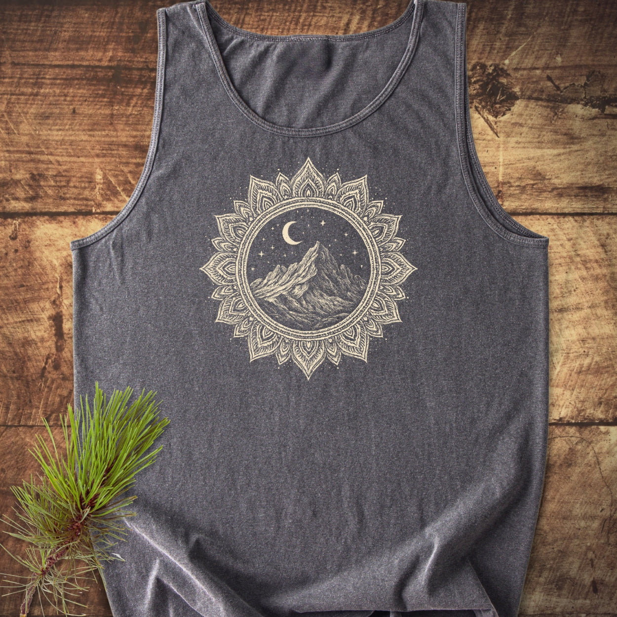 Mountain Mandala Tank Top featuring a cream mandala graphic with mountains, moon, and stars on charcoal-gray fabric, laid on wood beside a pine branch, highlighting its relaxed fit and sustainability.