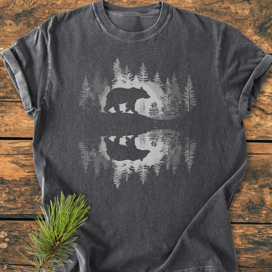 Bear Reflection - Vintage Wash Tee on wooden surface, featuring a bear and forest silhouette graphic with mirrored reflection. Styled with a pine bough, highlighting the tee's relaxed fit and soft-washed fabric.