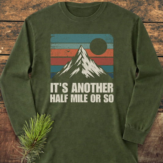 The It's Another Half Mile Or So Long-Sleeve Tee features a mountain graphic with retro stripes and text, on forest-green cotton, laid on wood with a pine sprig beside it.
