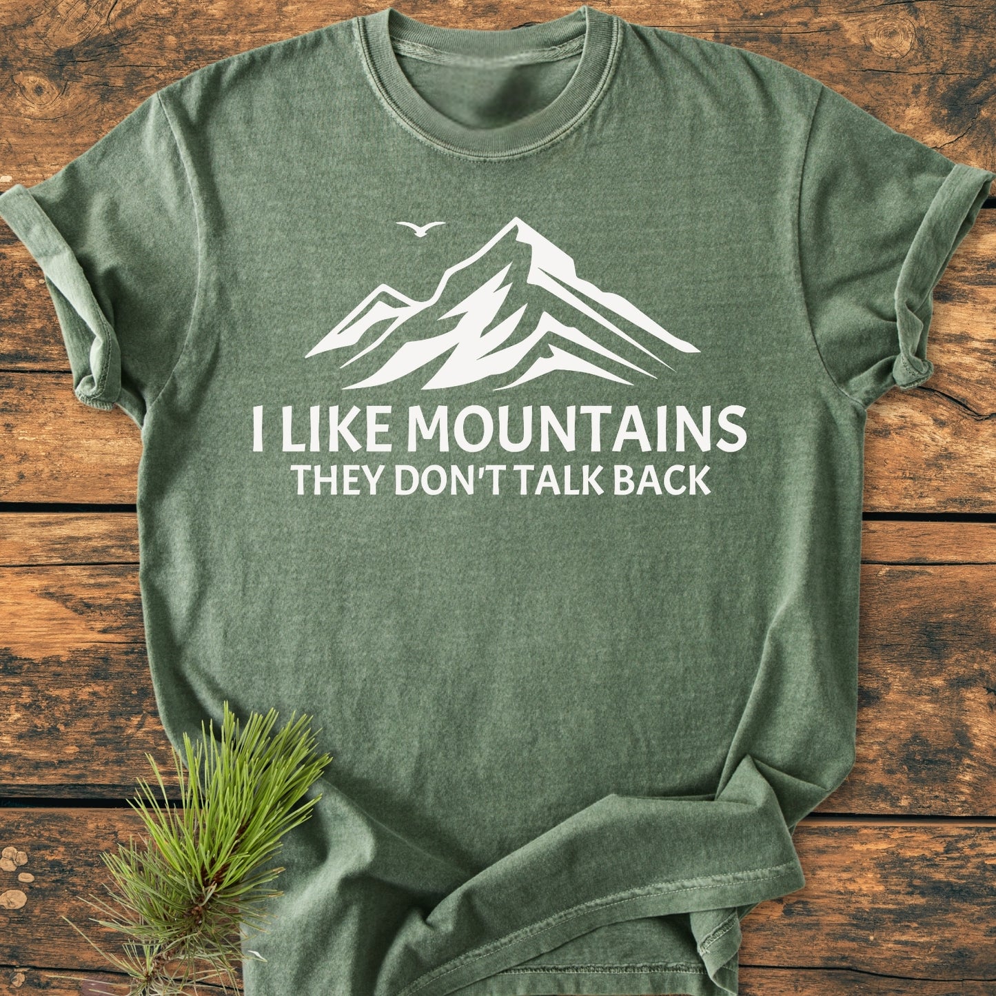 I Like Mountains - Vintage Wash Tee featuring a white stylized mountain print with slogan, laid flat on wood, complemented by a pine branch for a nature-inspired look.