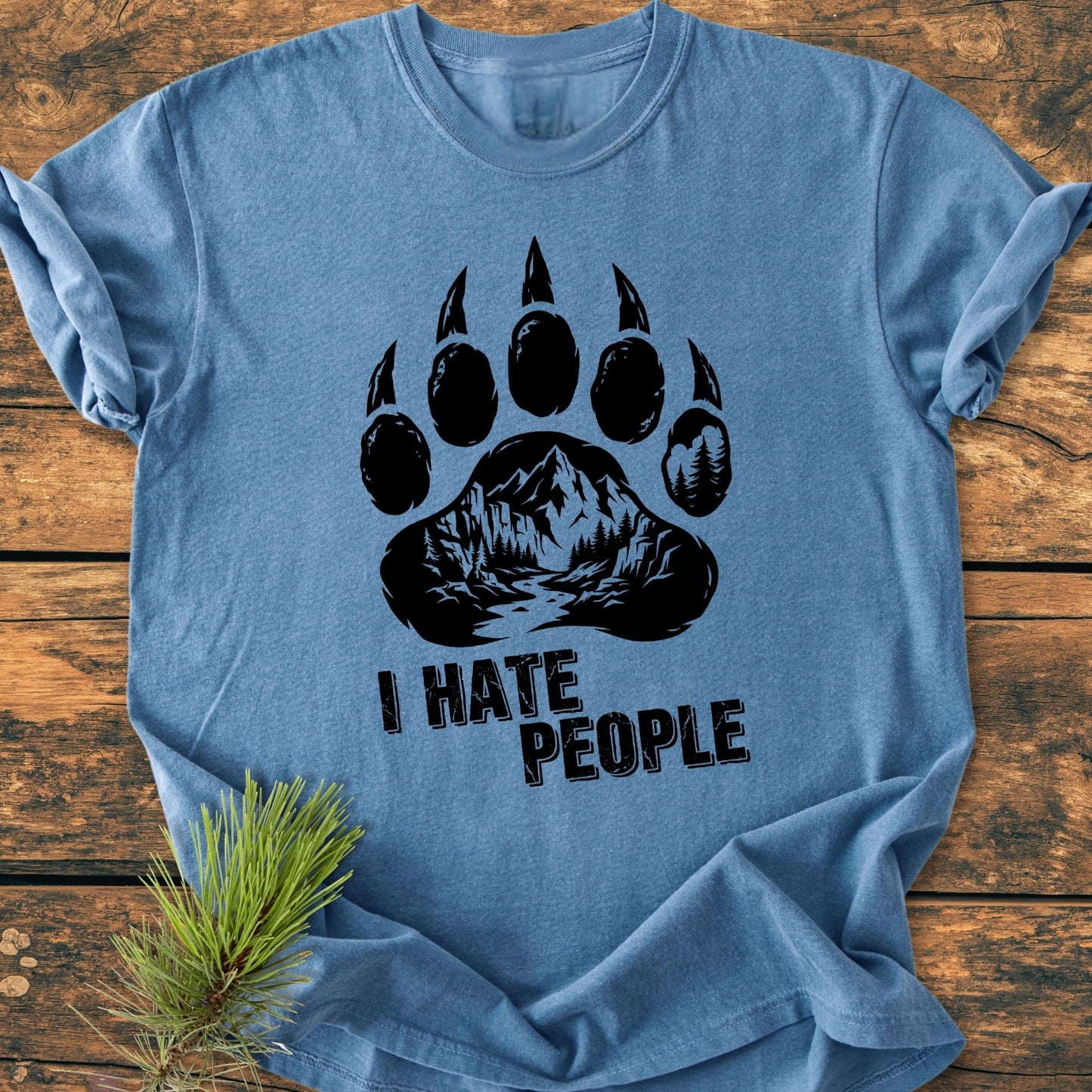 I Hate People - Vintage Wash Tee: Heather-blue shirt featuring a black stylized bear paw with mountain and forest design, text below. Laid on rustic wood with pine sprig beside.