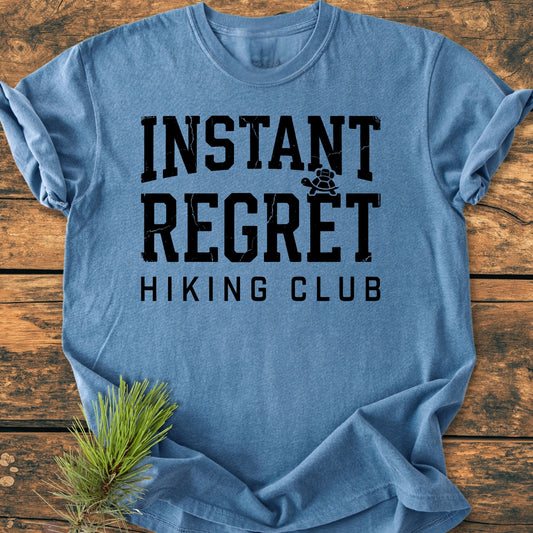 Instant Regret Hiking Club - Vintage Wash Tee features distressed black lettering and a turtle silhouette on a heathered-blue T-shirt, styled with pine needles on a wooden surface.