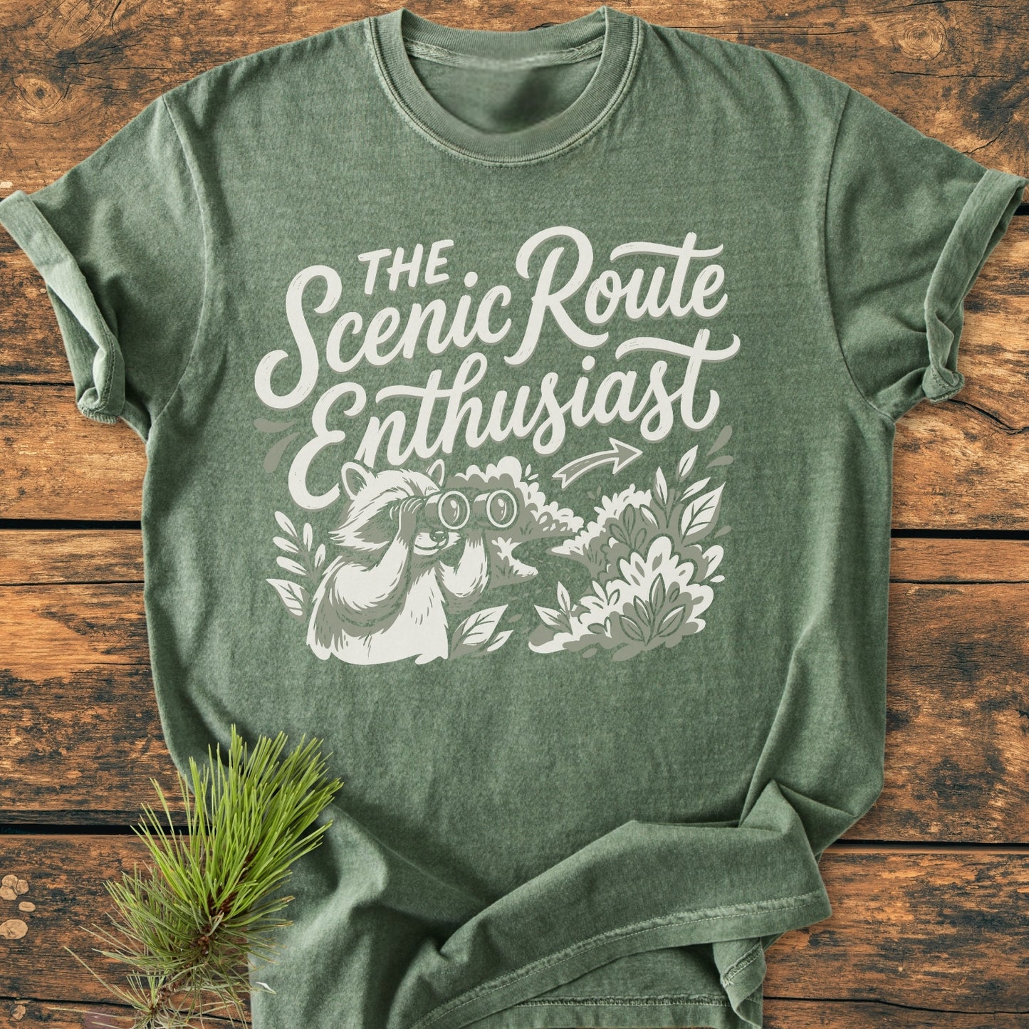 Scenic Route Raccoon - Vintage Wash Tee features a white slogan and raccoon graphic on forest-green fabric, laid on wood. Pine sprig accents its outdoorsy theme, showcasing a relaxed fit and durable stitching.