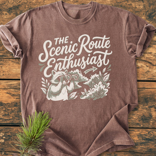 Scenic Route Raccoon - Vintage Wash Tee features a raccoon graphic with binoculars on an earth-tone shirt, laid flat on wood, styled with a pine sprig, highlighting its relaxed fit and cozy fabric.