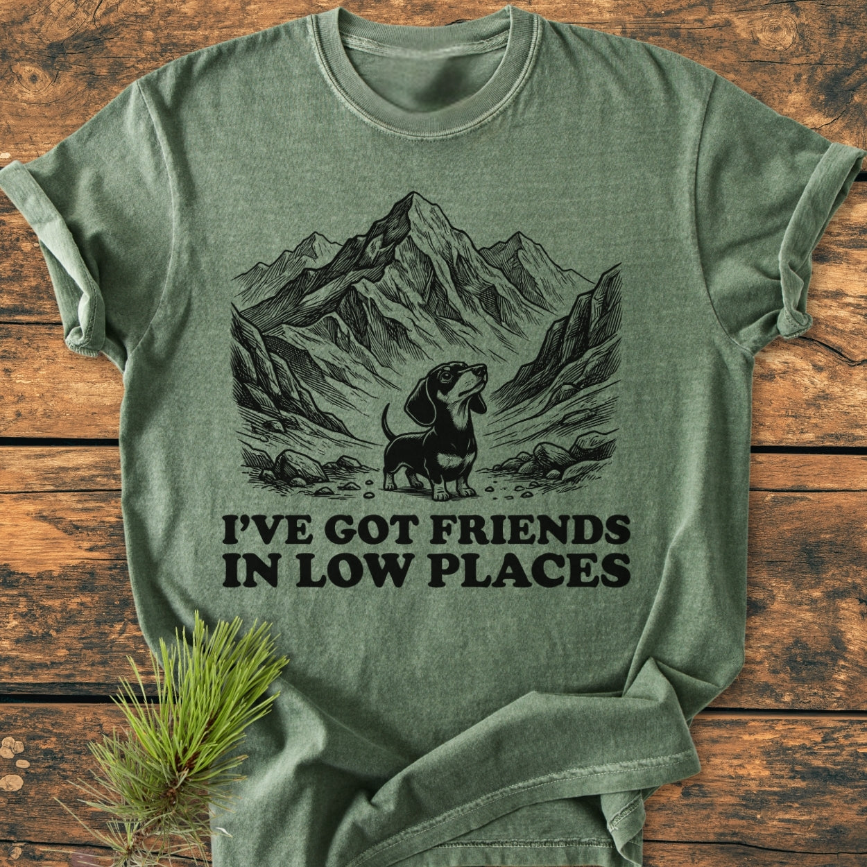 Friends In Low Places - Vintage Wash Tee features a black line drawing of a mountain valley with a dachshund and the slogan, perfect for a cozy, relaxed fit with durable construction.